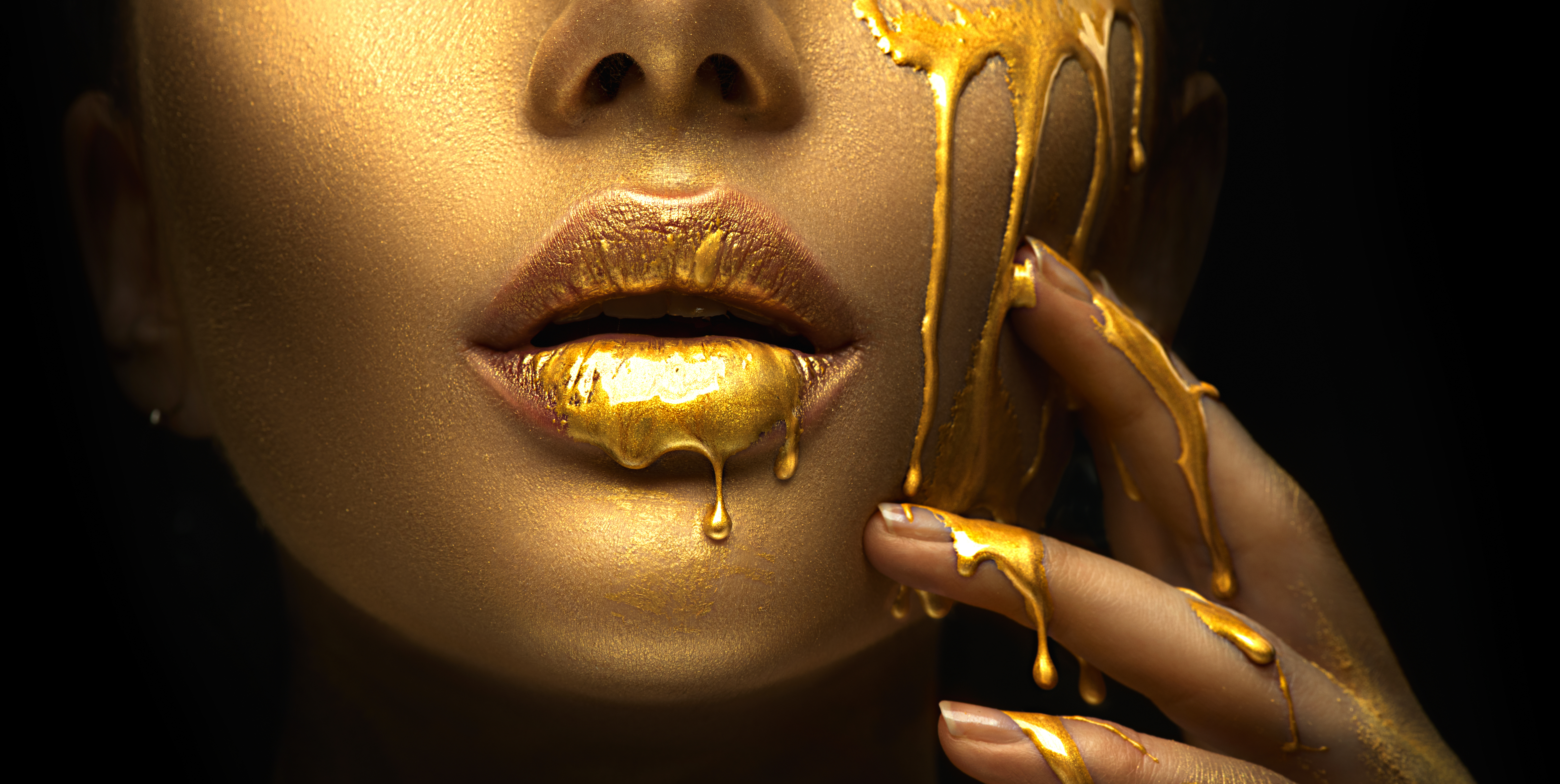 Golden paint smudges drips from the face lips and hand, golden liquid drops on beautiful model girl's mouth, creative abstract makeup. Beauty woman face